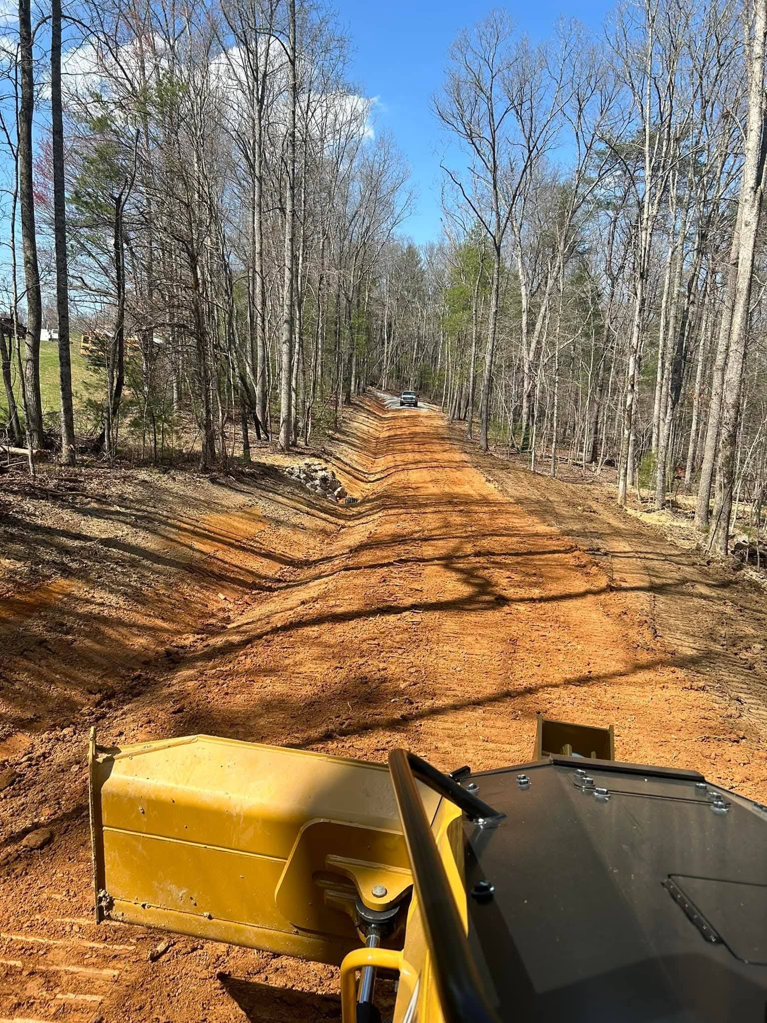 A view from the cab of a piece of yellow heavy machinery grading a dirt path through a wooded area on a sunny day.