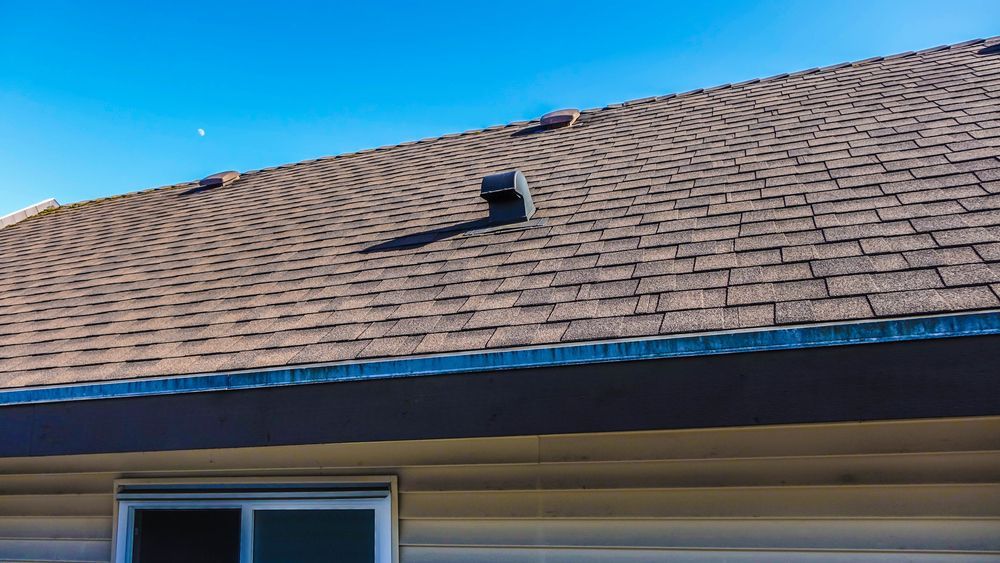 Brown asphalt shingle roof with a vent pipe against a blue sky. The roof sits above beige siding and a window.