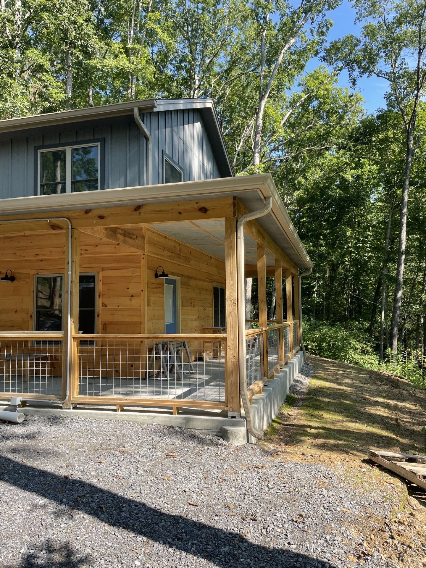 Cabin with a wrap-around porch in a wooded area. Light brown wood siding and grey roof. Gravel driveway.