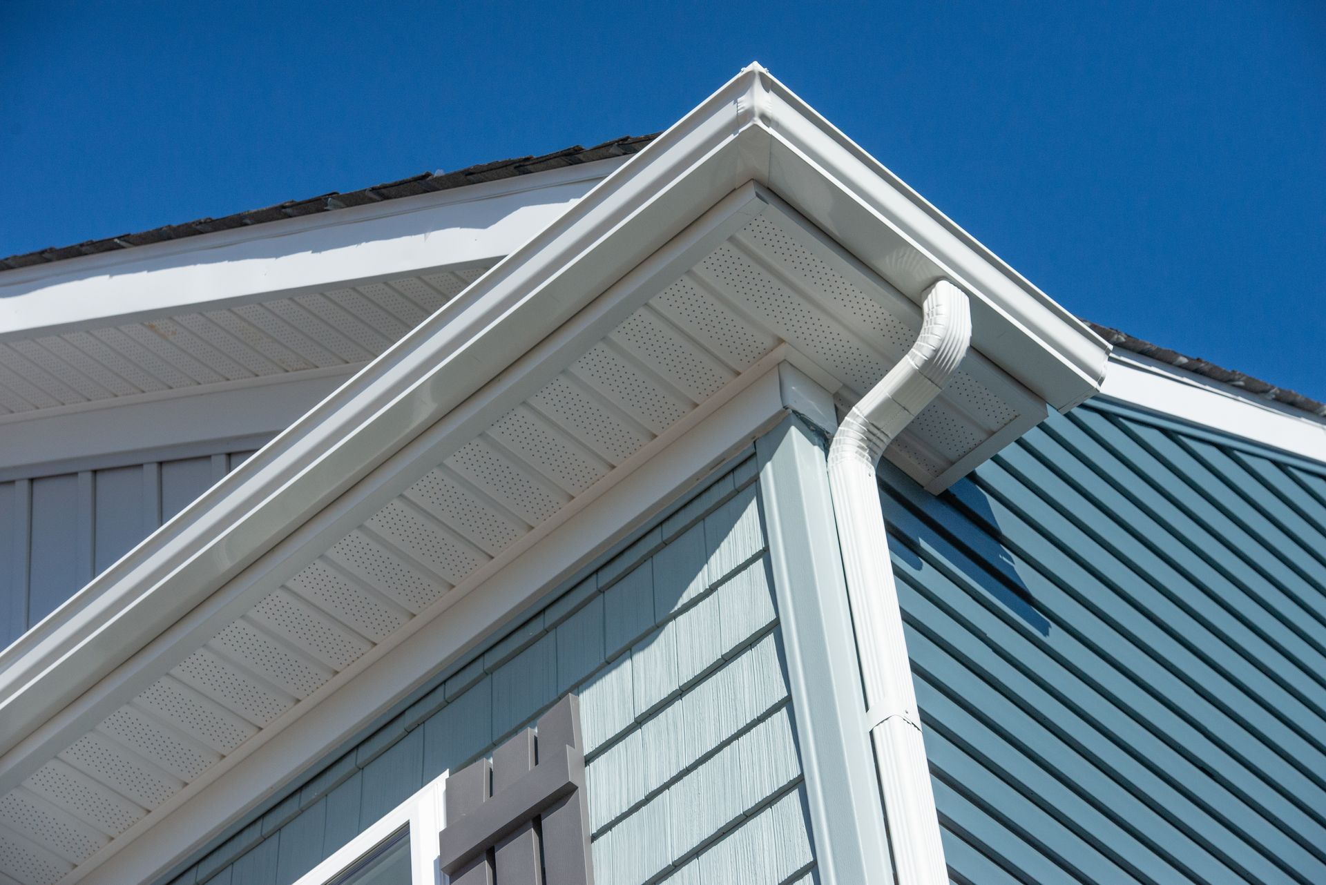 Corner of a house with white trim, blue siding, and a gutter against a clear, blue sky.
