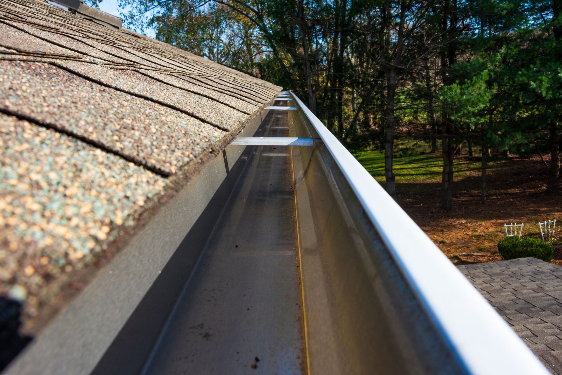 Gutter on a roof, viewed from above, with brown shingles and a backdrop of green trees.