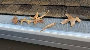 Gloved hand installing a white gutter guard on a roof's edge, over a black gutter. Green grass in background.