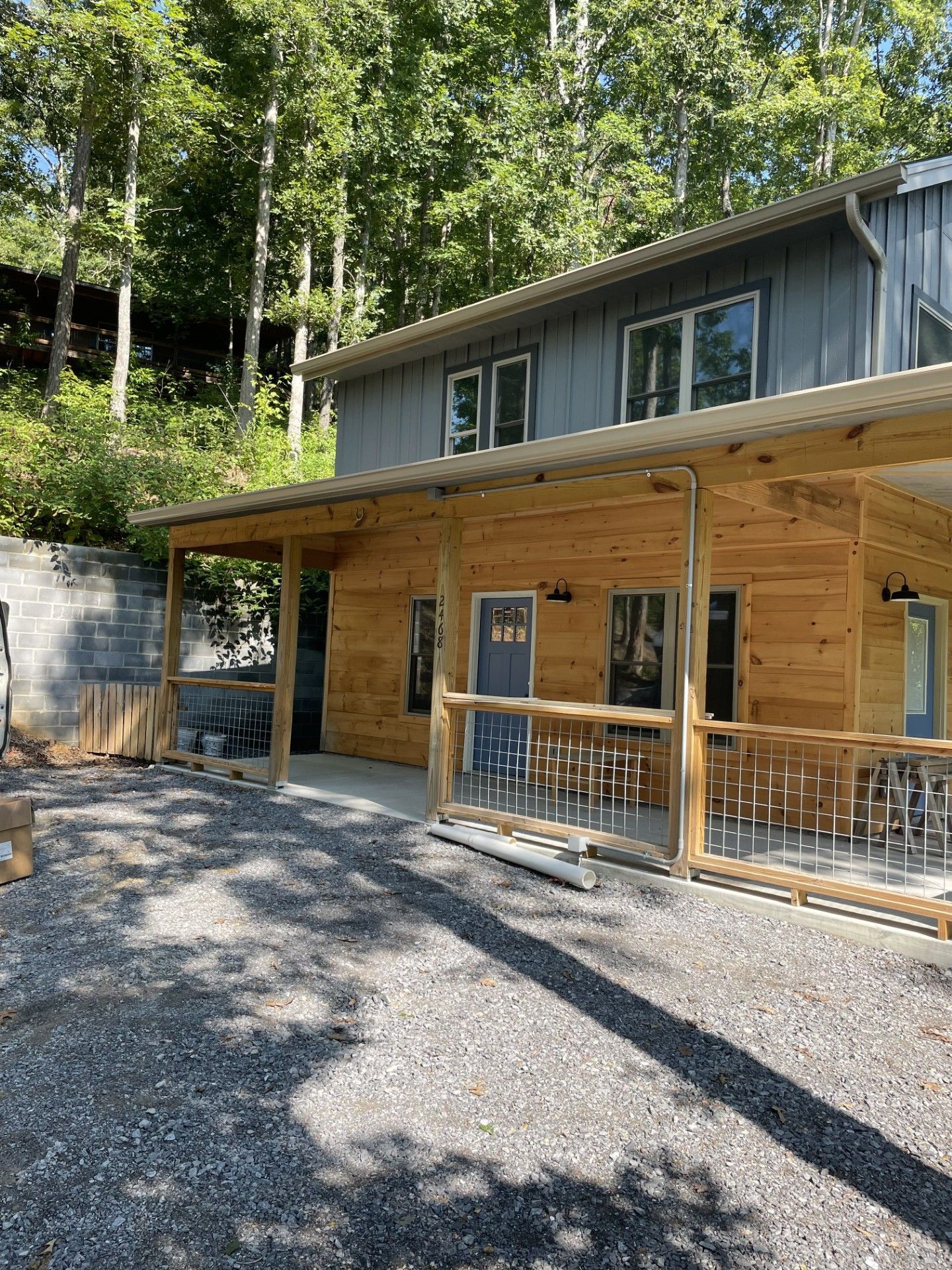 Two-story cabin with a wraparound porch and gravel driveway. Wooden exterior and gray roof, surrounded by trees.