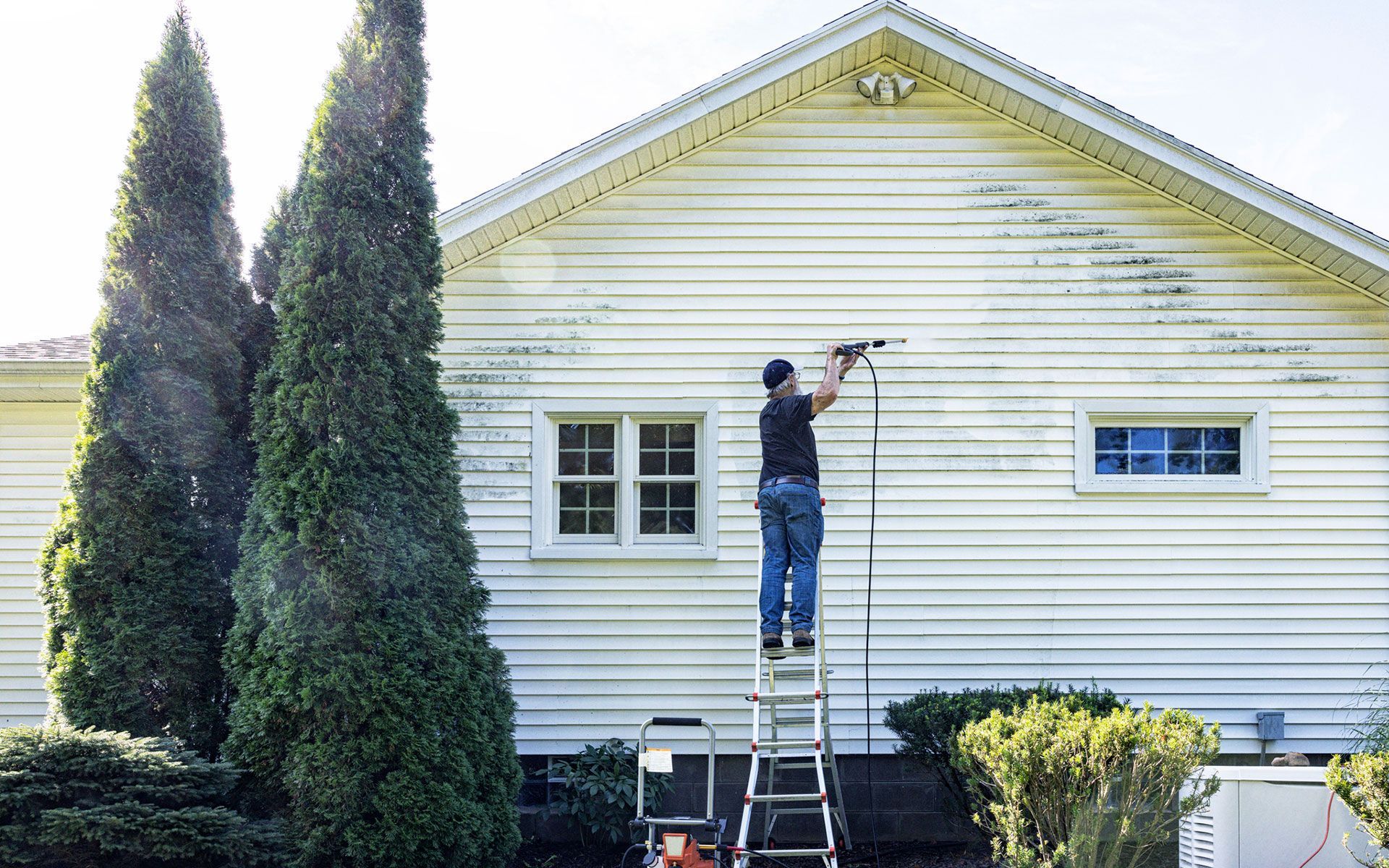 A man is standing on a ladder painting the side of a white house.