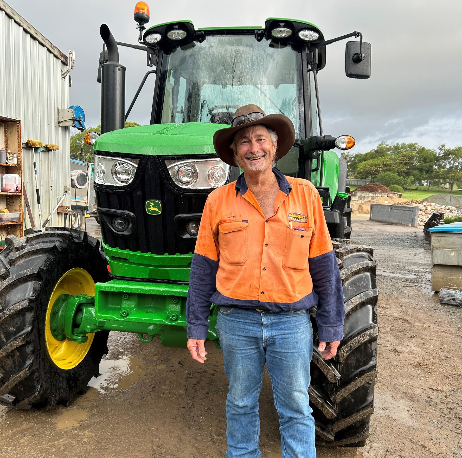 Man in work clothes smiles beside a green John Deere tractor in front of a building.