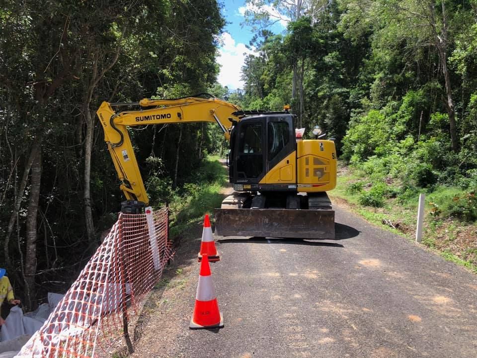 A Yellow Excavator Is Driving Down A Road Next To A Fence And Cones — Campbell's Truck and Bobcat and Landscape Supplies In Gympie, QLD