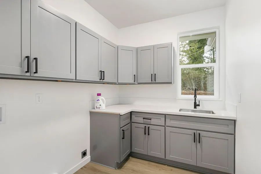 A laundry room with gray cabinets , a sink , and a window.