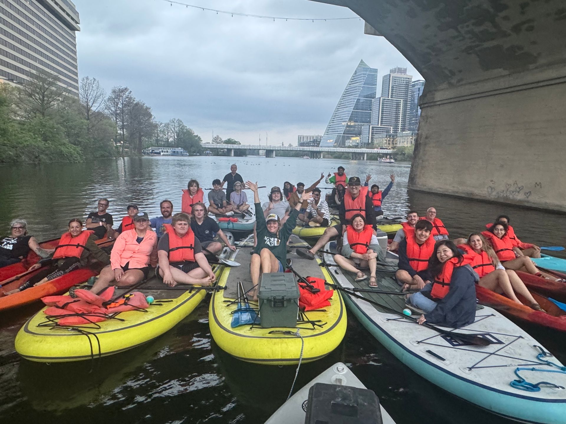 A group of people are sitting on rafts in the water under a bridge in the Austin skyline.