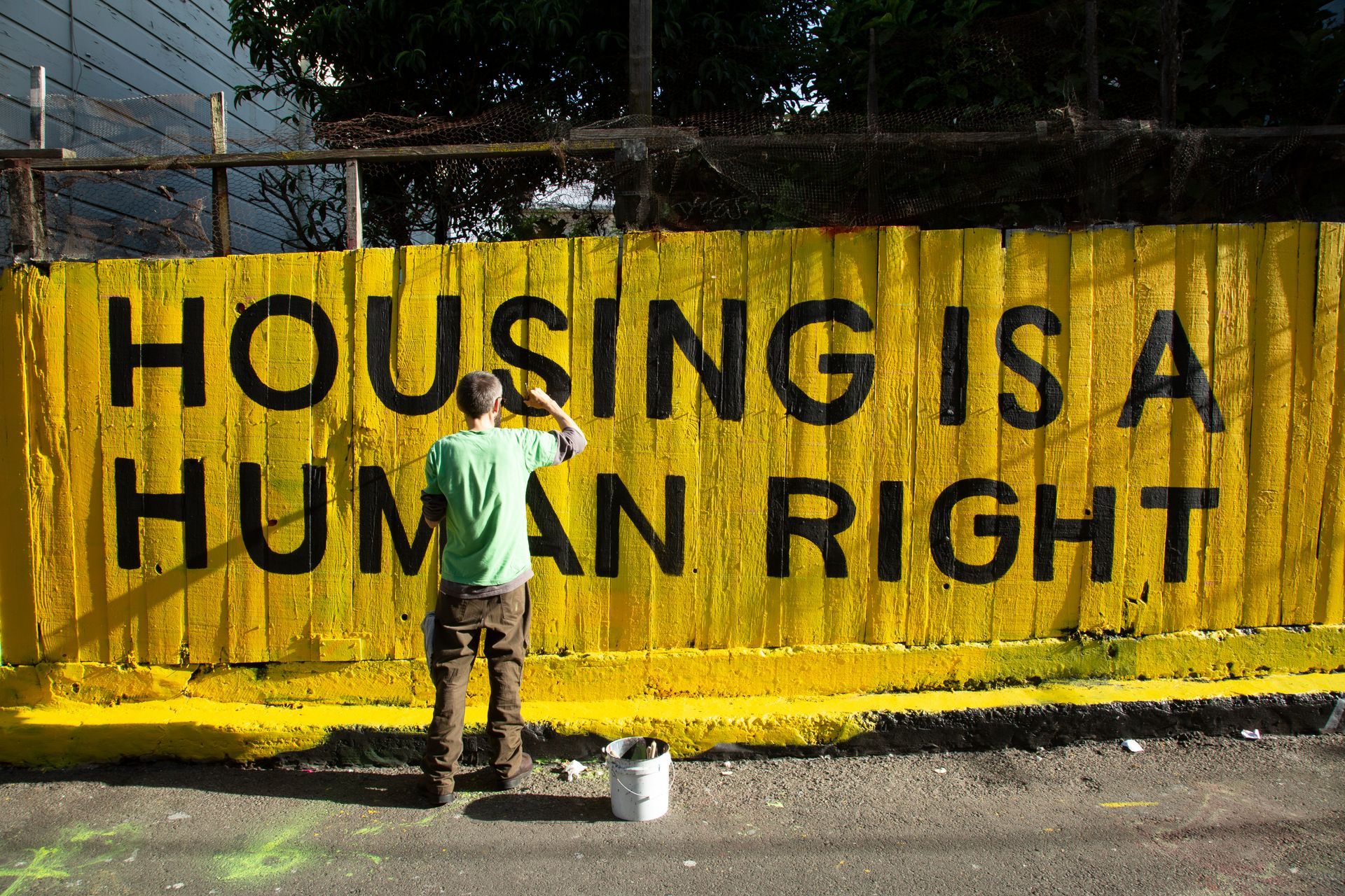 A man paints a yellow fence that says housing is a human right