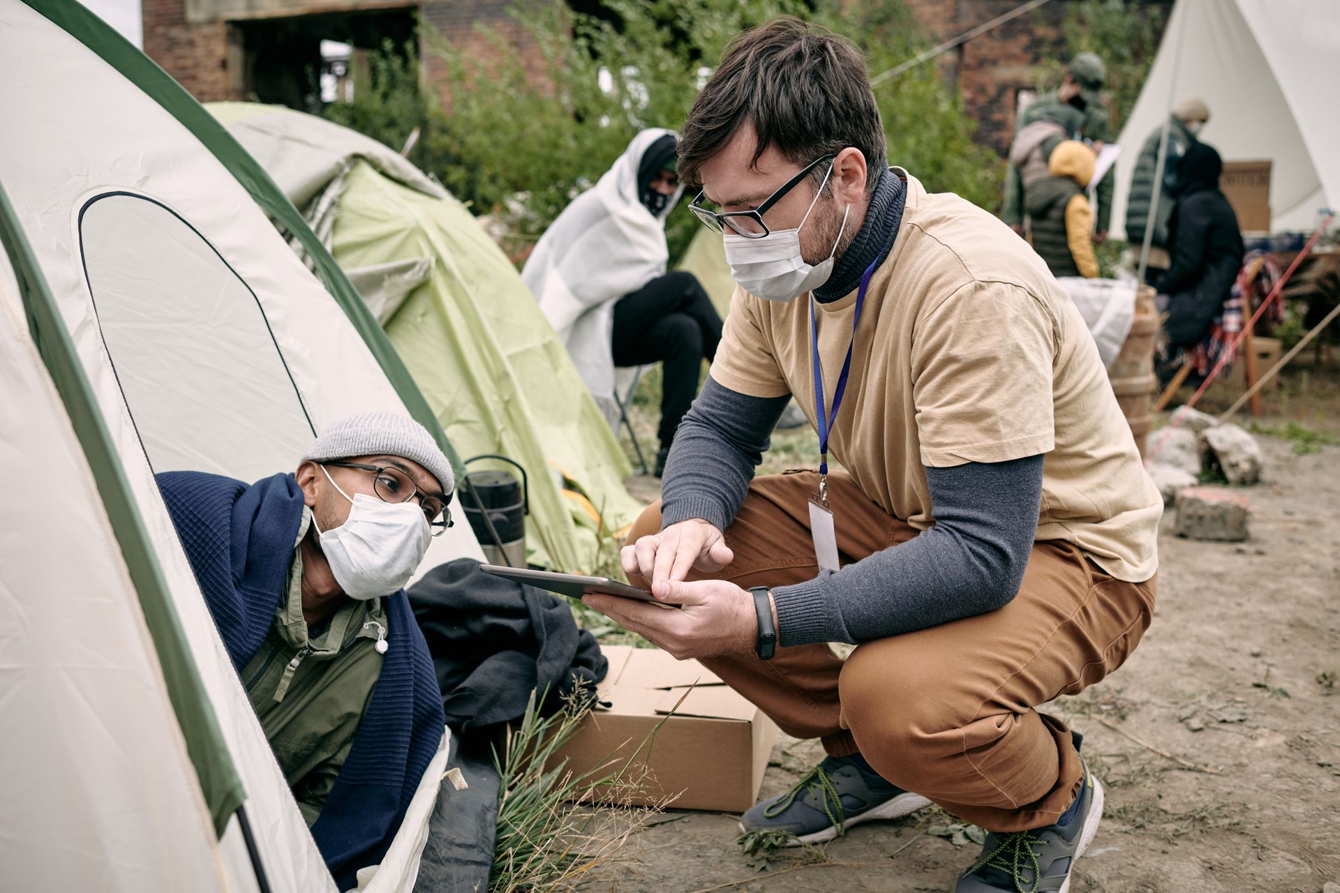 A man wearing a mask is kneeling down next to a tent and looking at a tablet.
