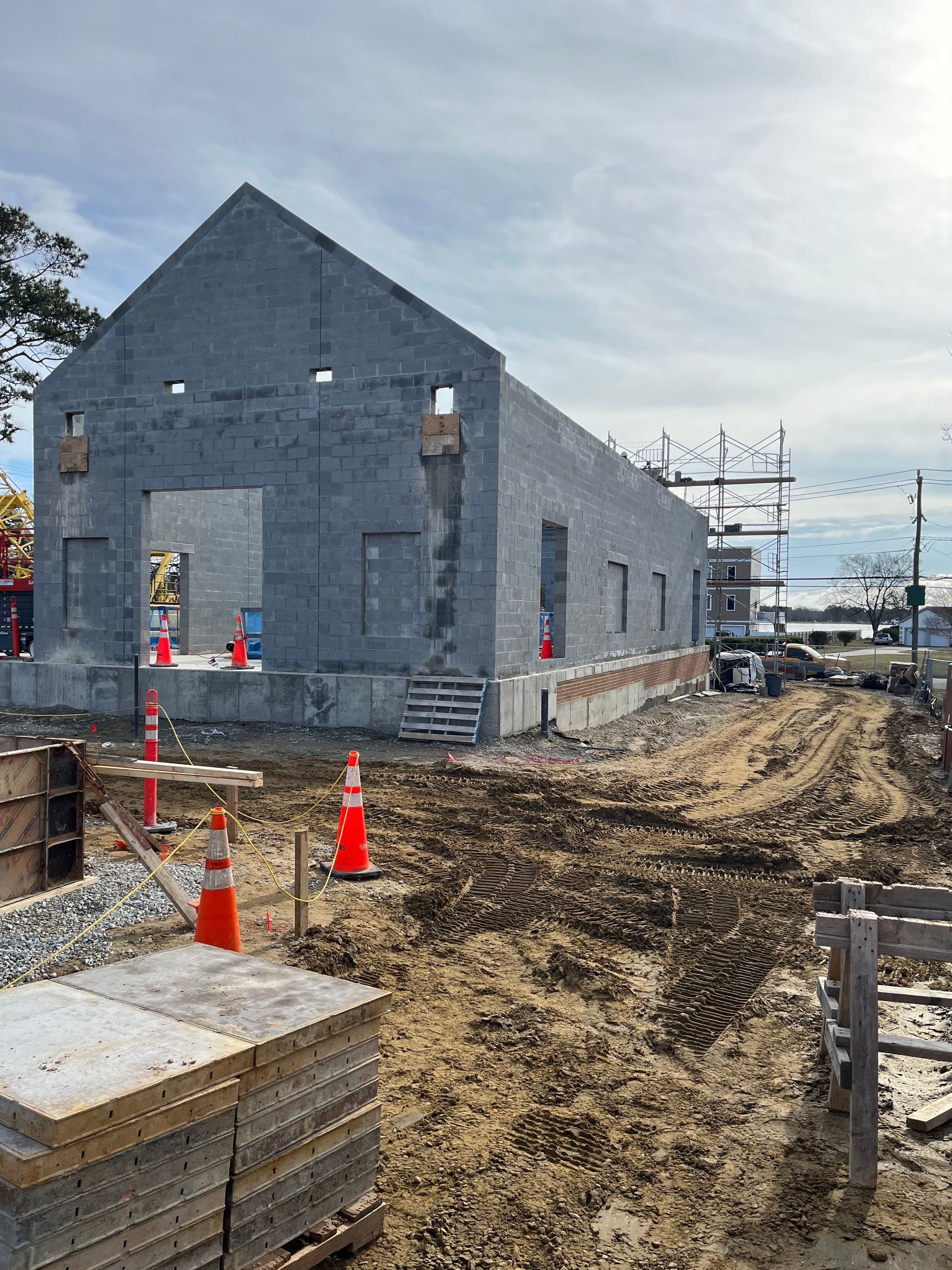 Gray industrial building under construction beside a muddy trench, with orange cones and stacked materials