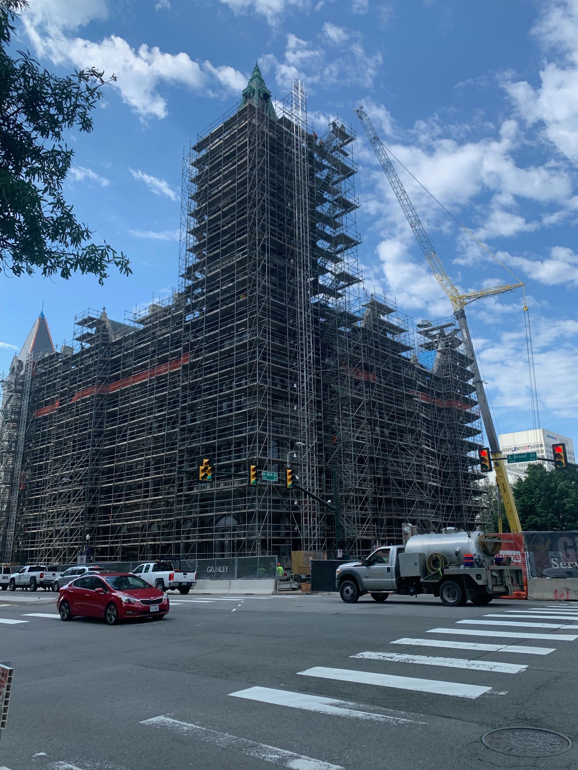 Scaffolding-covered building under construction at a city intersection with cars and a crane under a blue sky