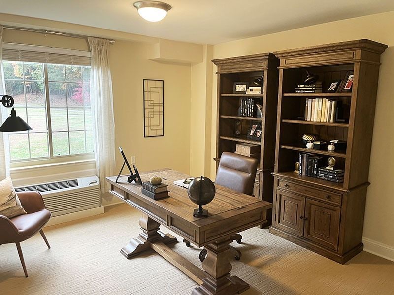 Home office with wooden desk, bookcases, and a window by a cream wall and carpeted floor