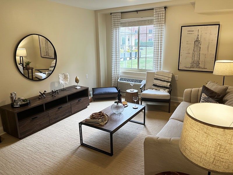Bright living room with beige sofa, coffee table, round mirror, and large window with blinds.