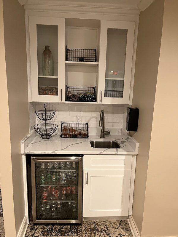 Compact kitchenette with white cabinets, stainless sink, small fridge, and glass-front wall shelves.