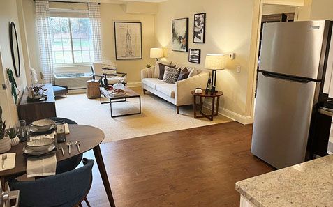 Open-plan living room with beige sofa, coffee table, and stainless steel refrigerator beside a kitchen counter.