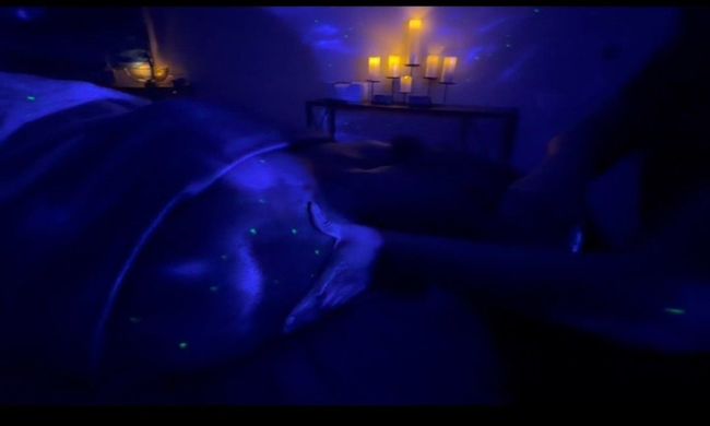 Blue-lit room with a person reclining in bed and a glowing candle centerpiece in the background