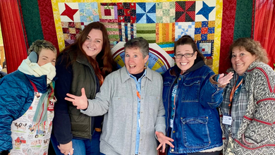 five women wearing warm clothes standing in front of the NLHG quilt