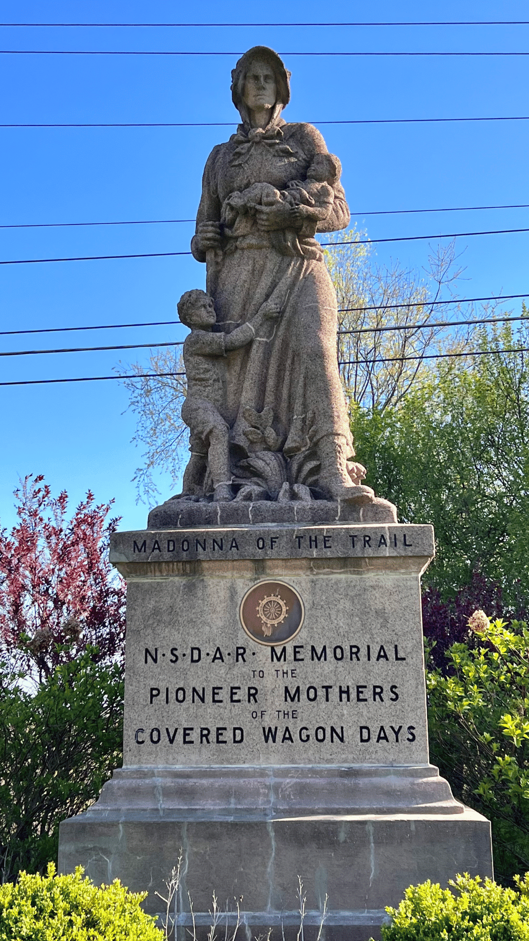 statue of a woman holding a baby with a child holding her skirt with the words Madonna of the Trail NSDAR Memorial Pioneer to the Mothers of the Covered Wagon Days underneath