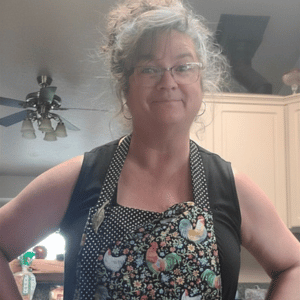 Smiling woman in a patterned apron and glasses, standing in a kitchen with hands on hips.