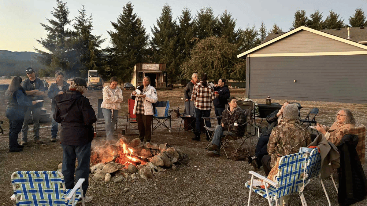 Women around a bonfire outdoors near a building and trees; some are standing, some are seated.