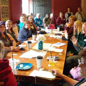 A group of people sit around a long wooden conference table in a meeting, with papers and beverages spread out.