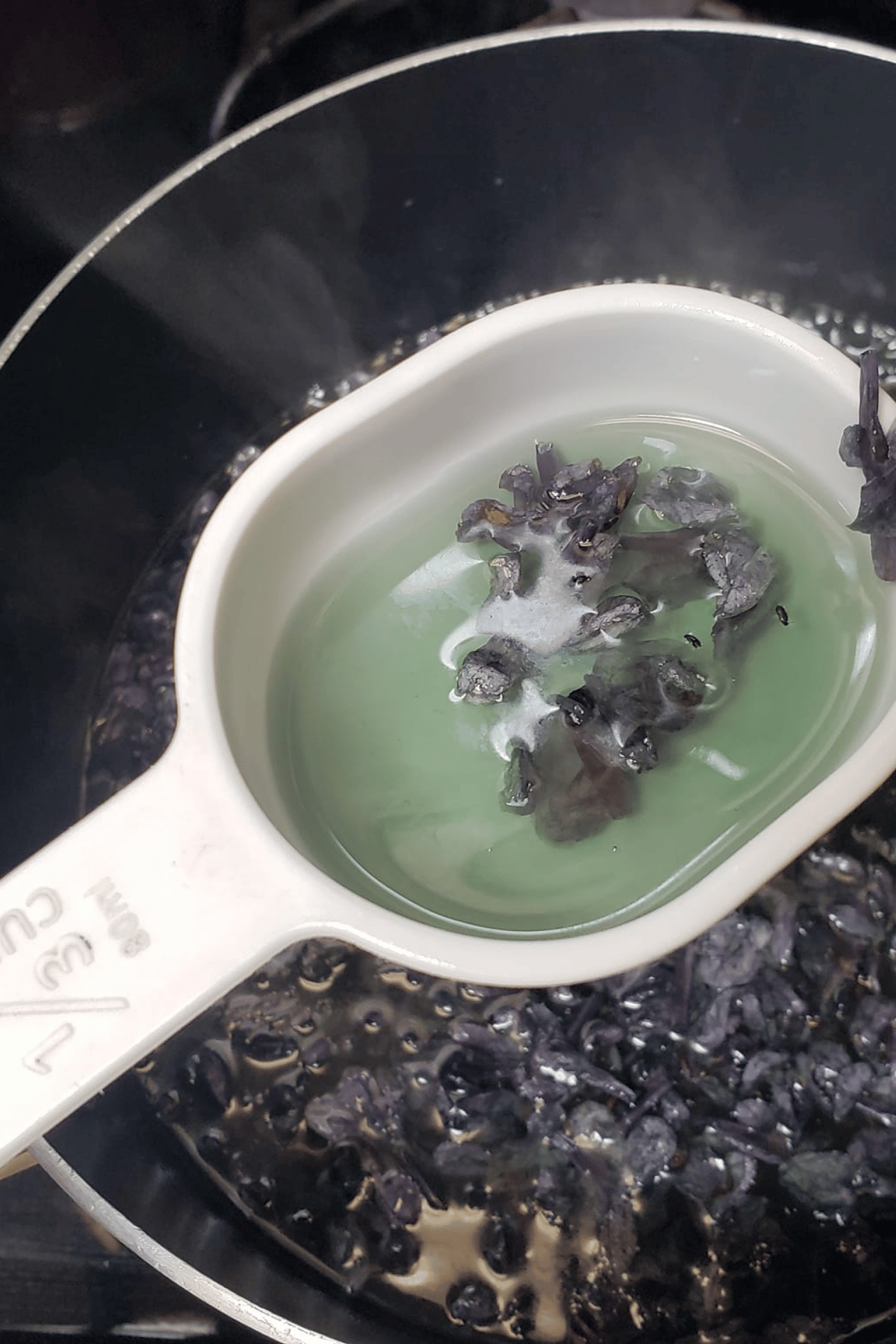 A white measuring cup filled with light green liquid and dark lilac petals in the cup and pot