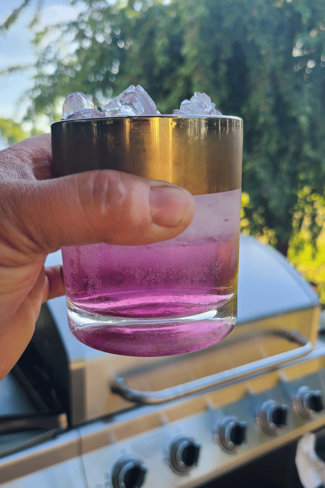 A hand holds a gold-rimmed glass filled with purple liquid and ice in front of an outdoor grill.