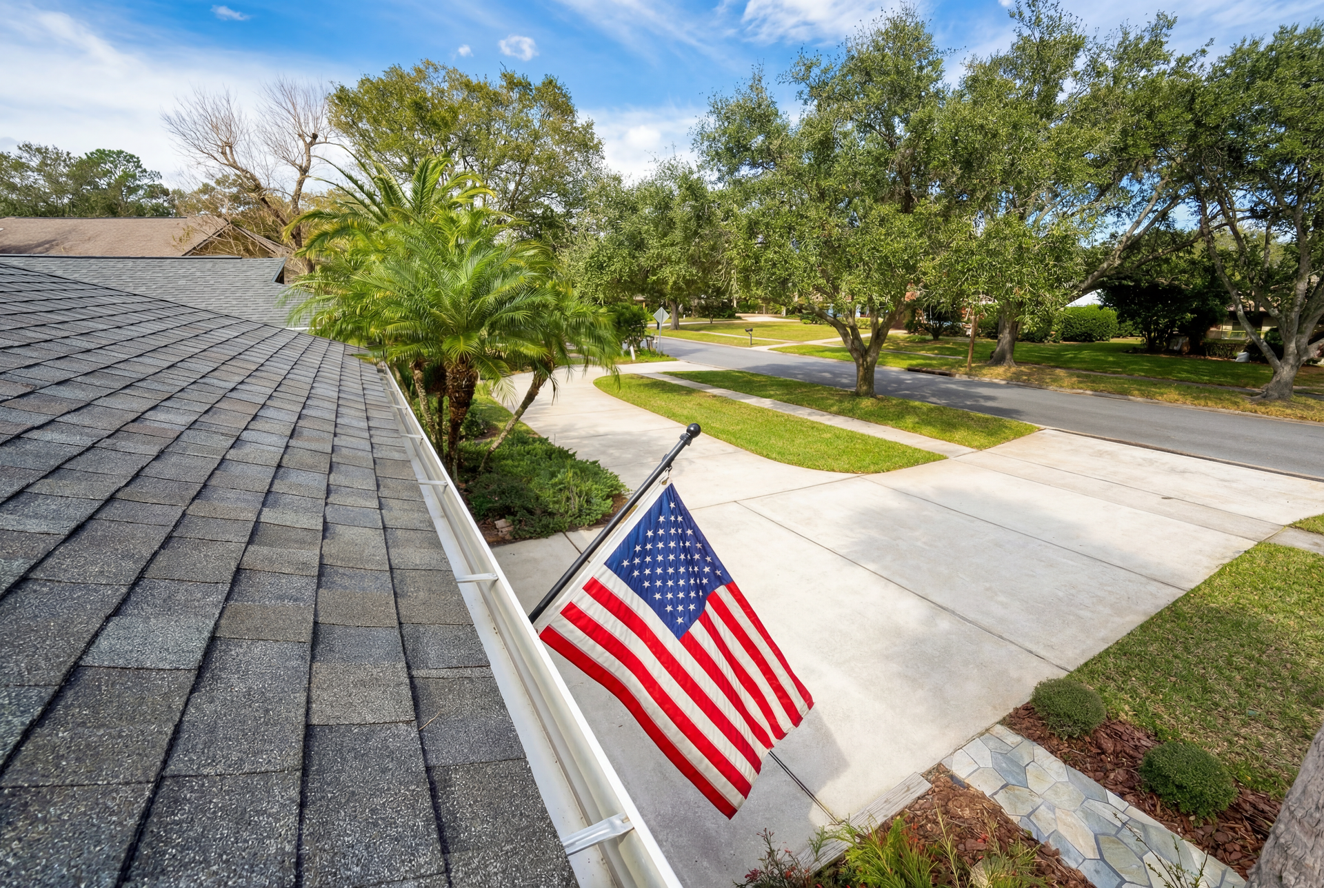 American flag flying in front of a house, driveway, and street, with trees and a roof in view.