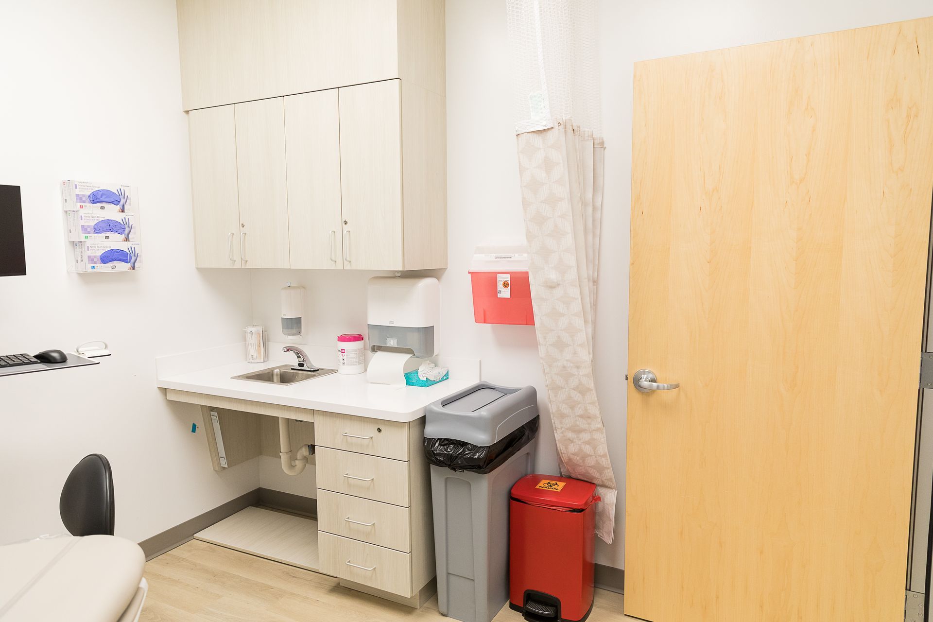 A doctor 's office with a sink , trash can , and chair.