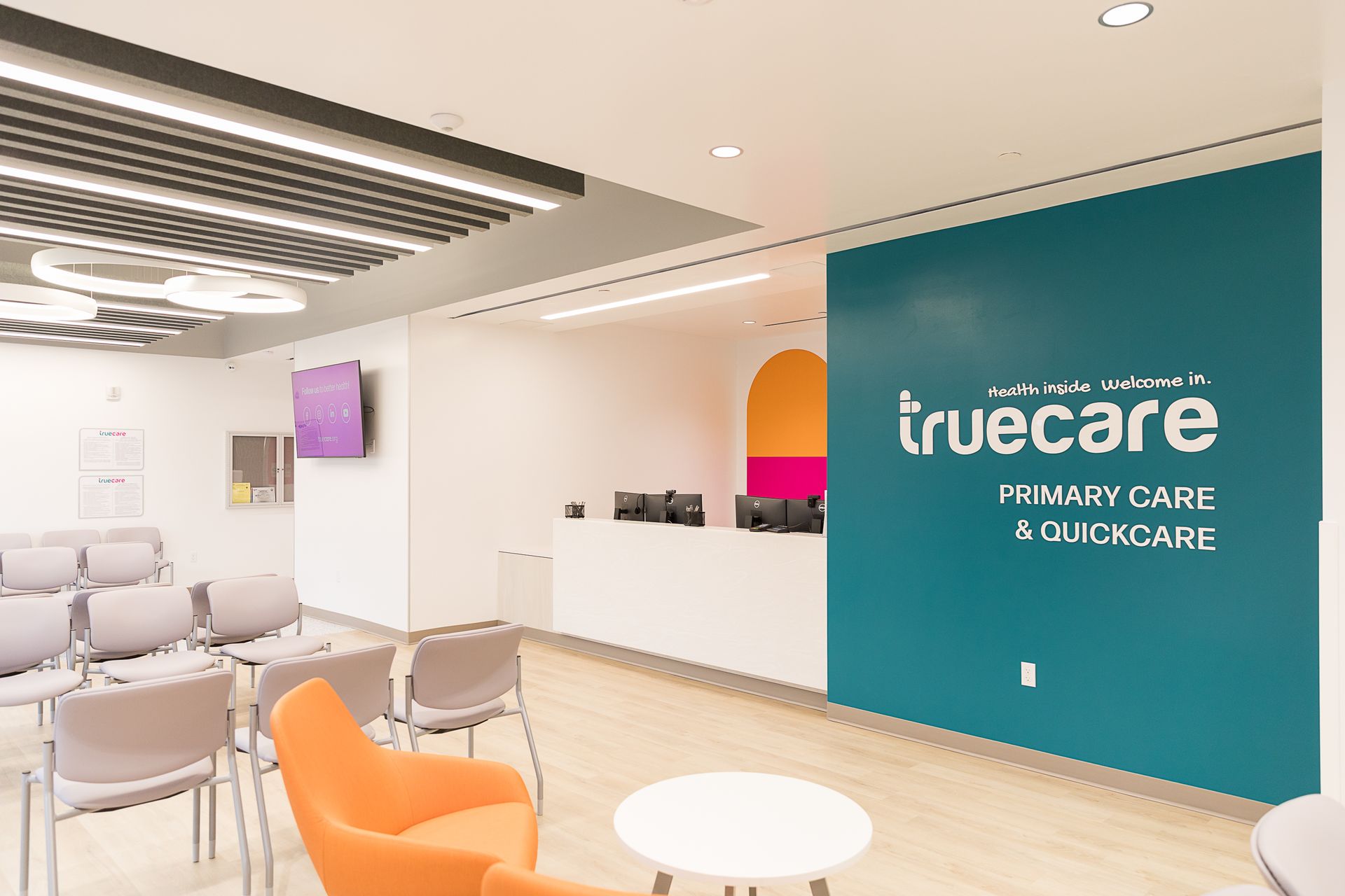 Interior view of a Truecare primary care office. Teal accent wall, white reception desk, orange chair, and seating.