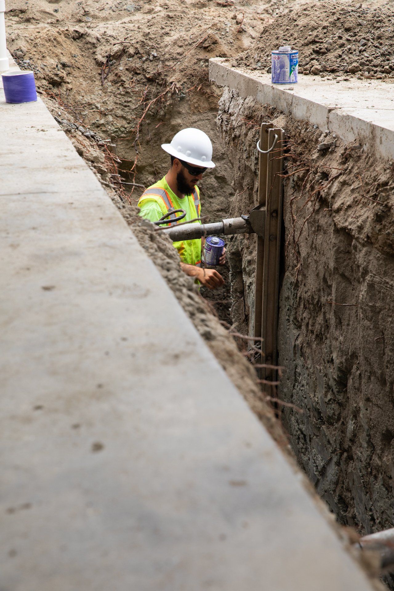 A construction worker wearing a hard hat and safety vest is working in a trench.