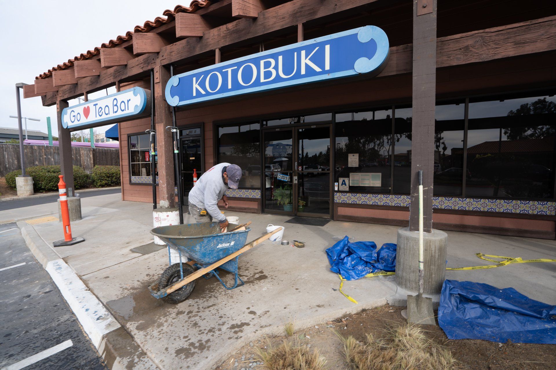 A man is pushing a wheelbarrow in front of a building that says kotobuki