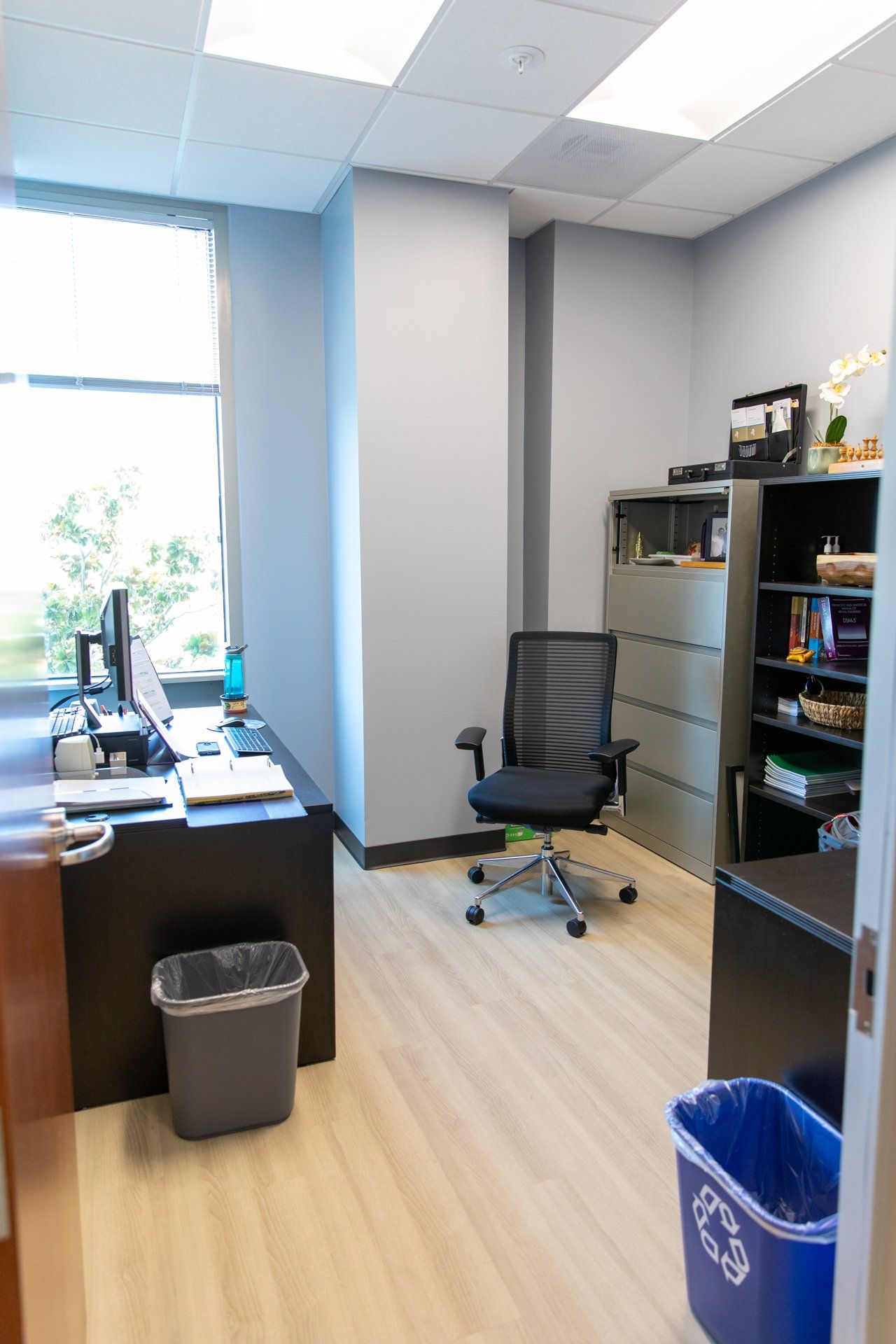 A small office with a desk , chair , trash can and a window.