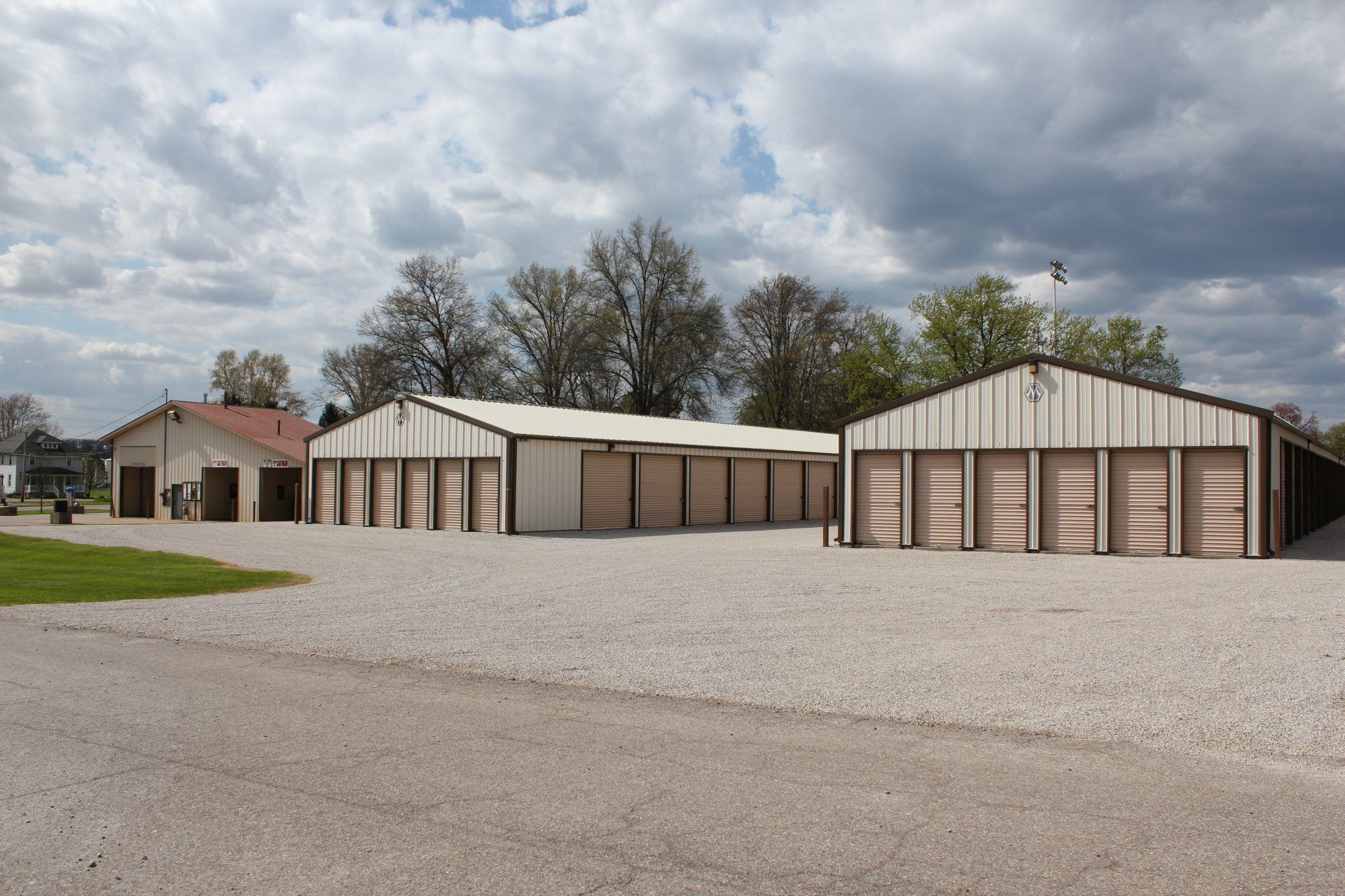 Storage units with brown roll-up doors, beige siding, and gravel parking under a cloudy sky.