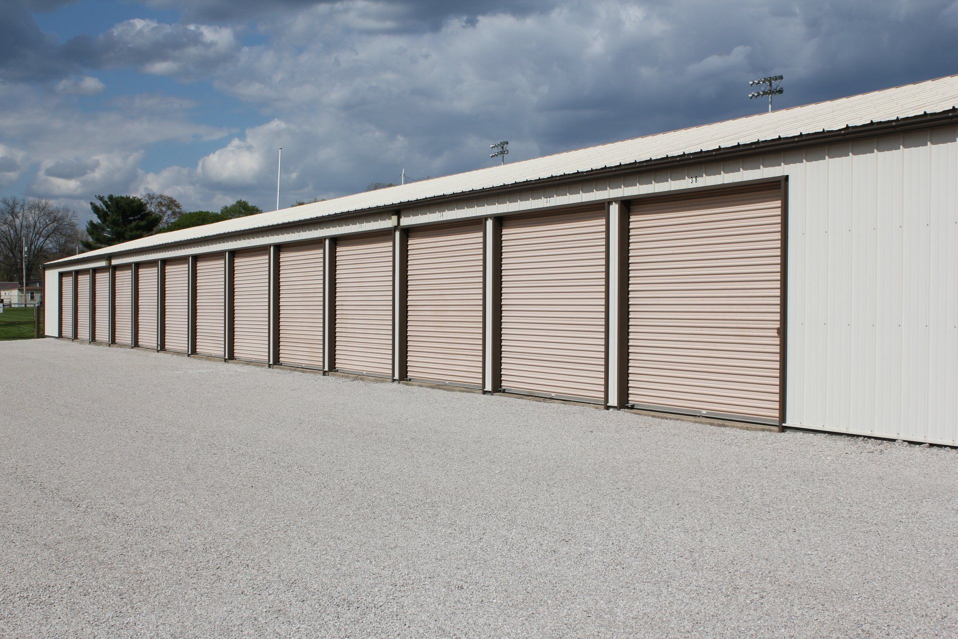 Storage units in a row, with beige roll-up doors, on a gravel lot under a cloudy sky.