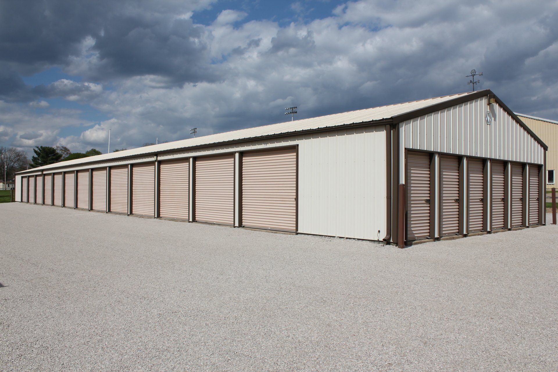 Storage units building with tan doors, brown trim, and a gravel parking area under a cloudy sky.