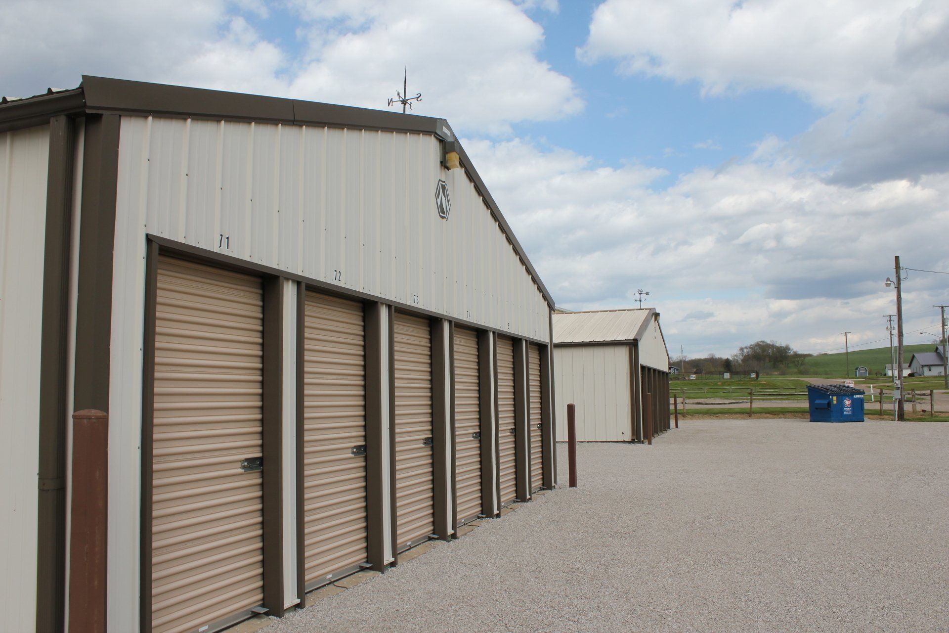Storage units with tan doors, brown trim, and a gravel lot under a cloudy sky.