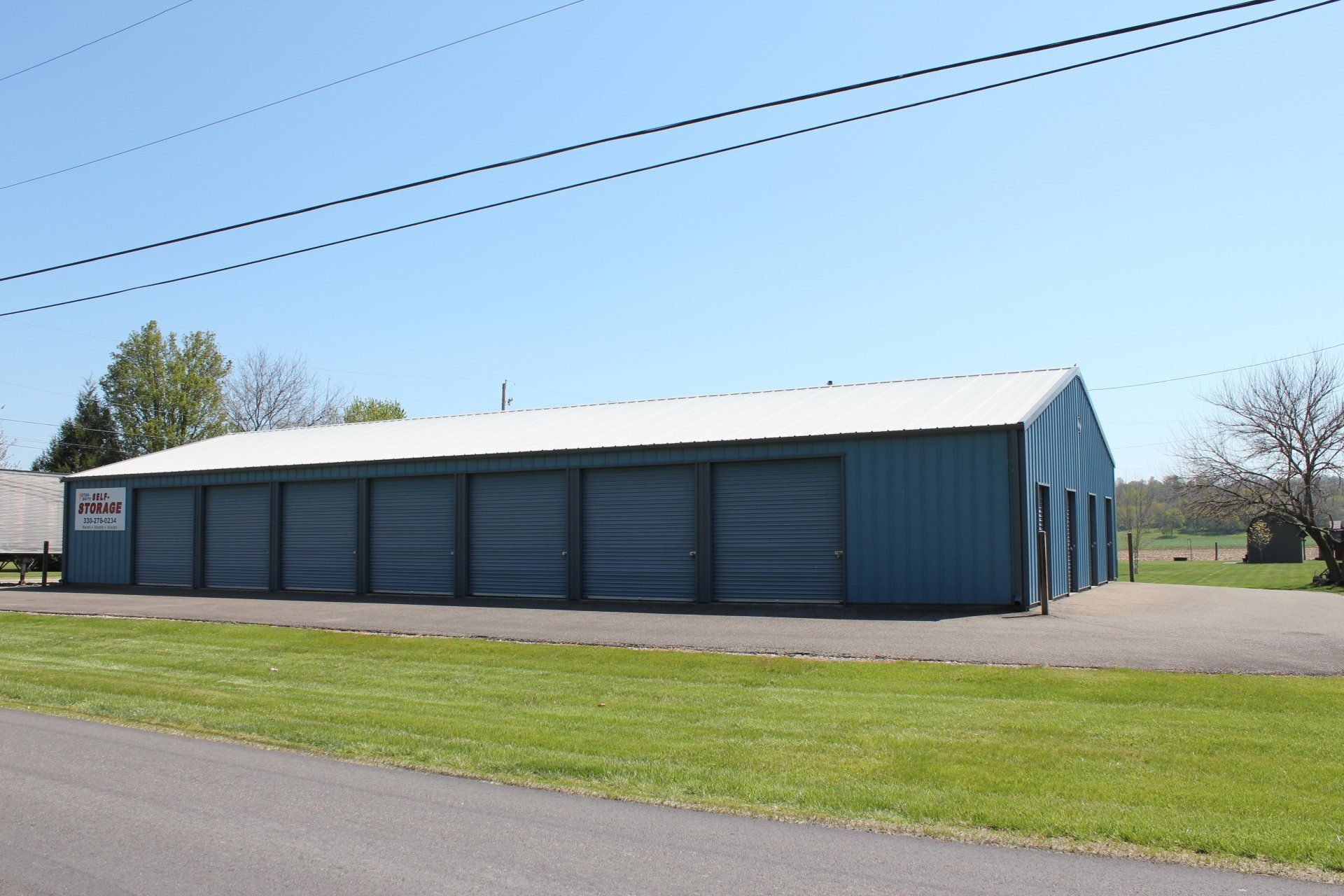 Blue storage units with metal roll-up doors, white roof, on a green lawn next to a road, under a blue sky.