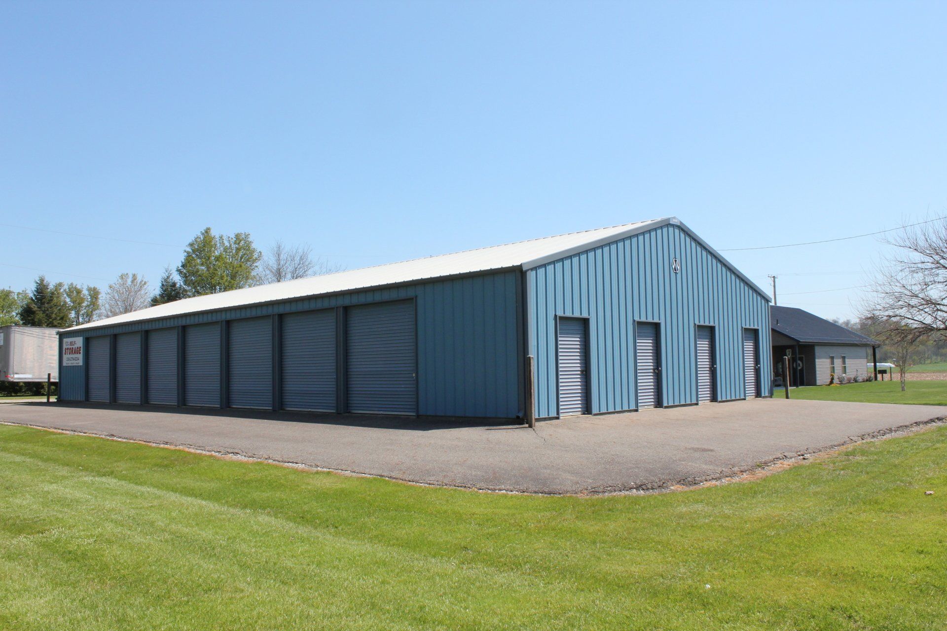 Blue storage facility with multiple roll-up doors and a white roof, situated on a concrete pad with grass in the foreground.