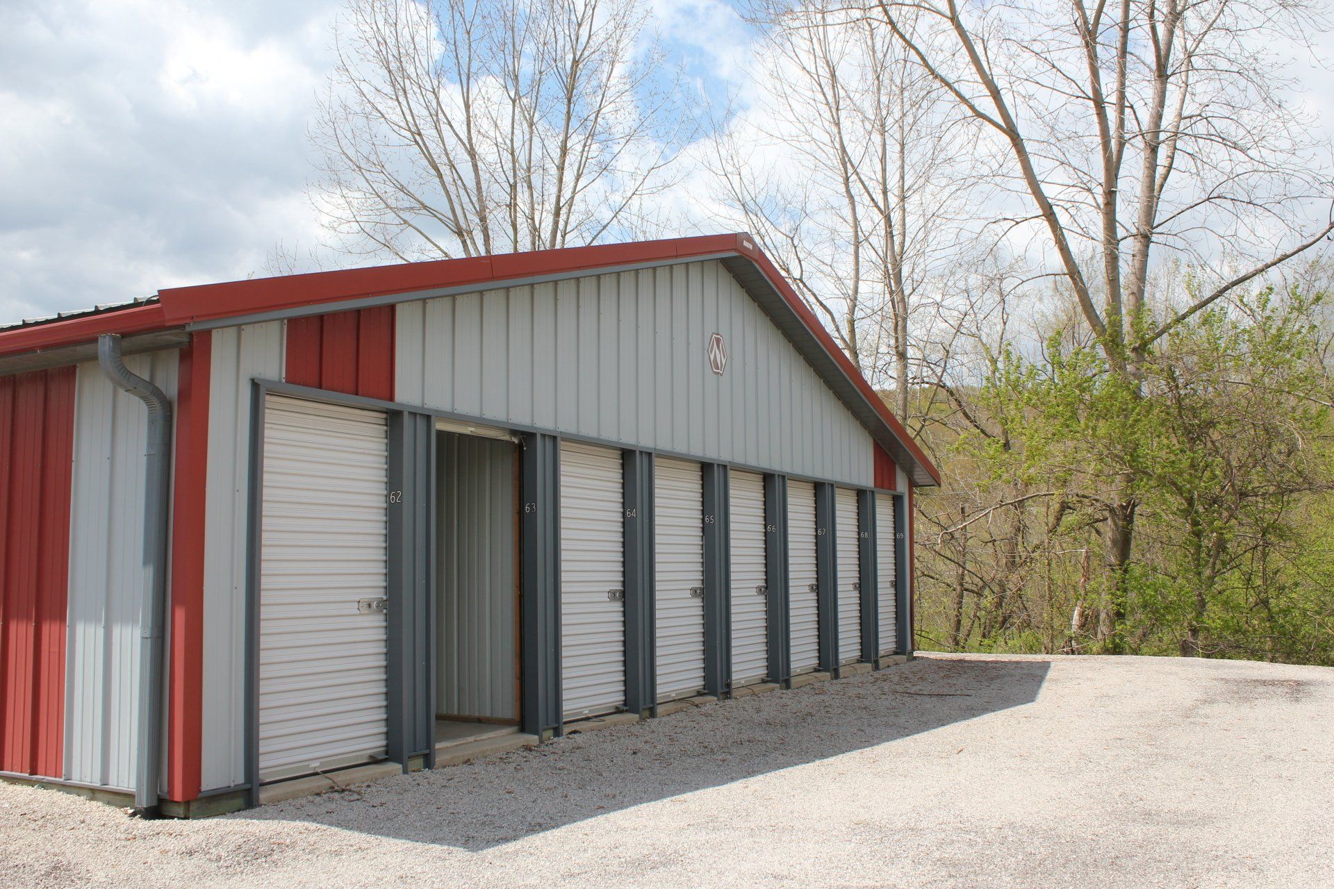 Storage units with red and gray exterior, gravel driveway, surrounded by trees under a cloudy sky.