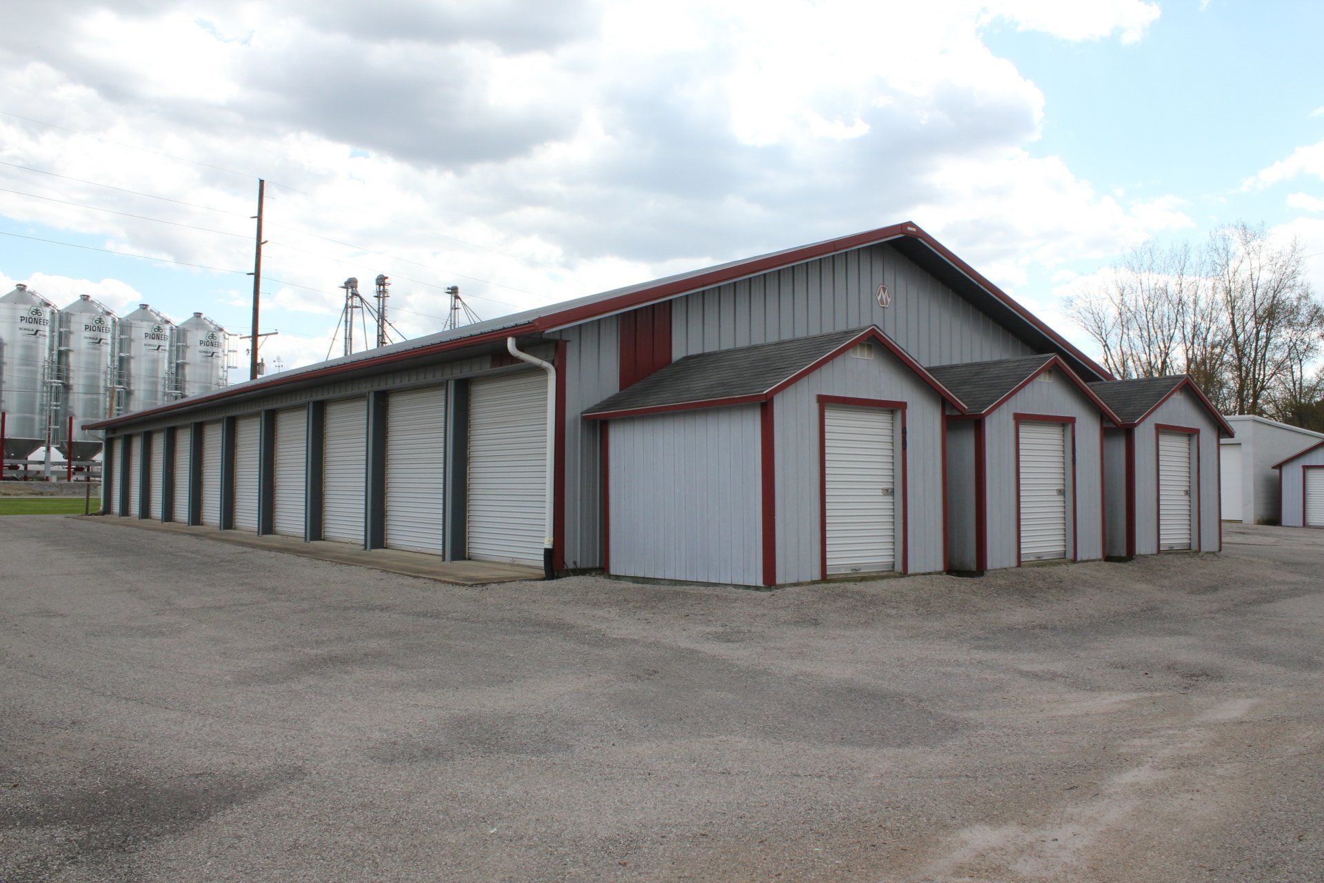 Gray storage units with red trim against a cloudy sky. Gravel ground. Silos in the background.