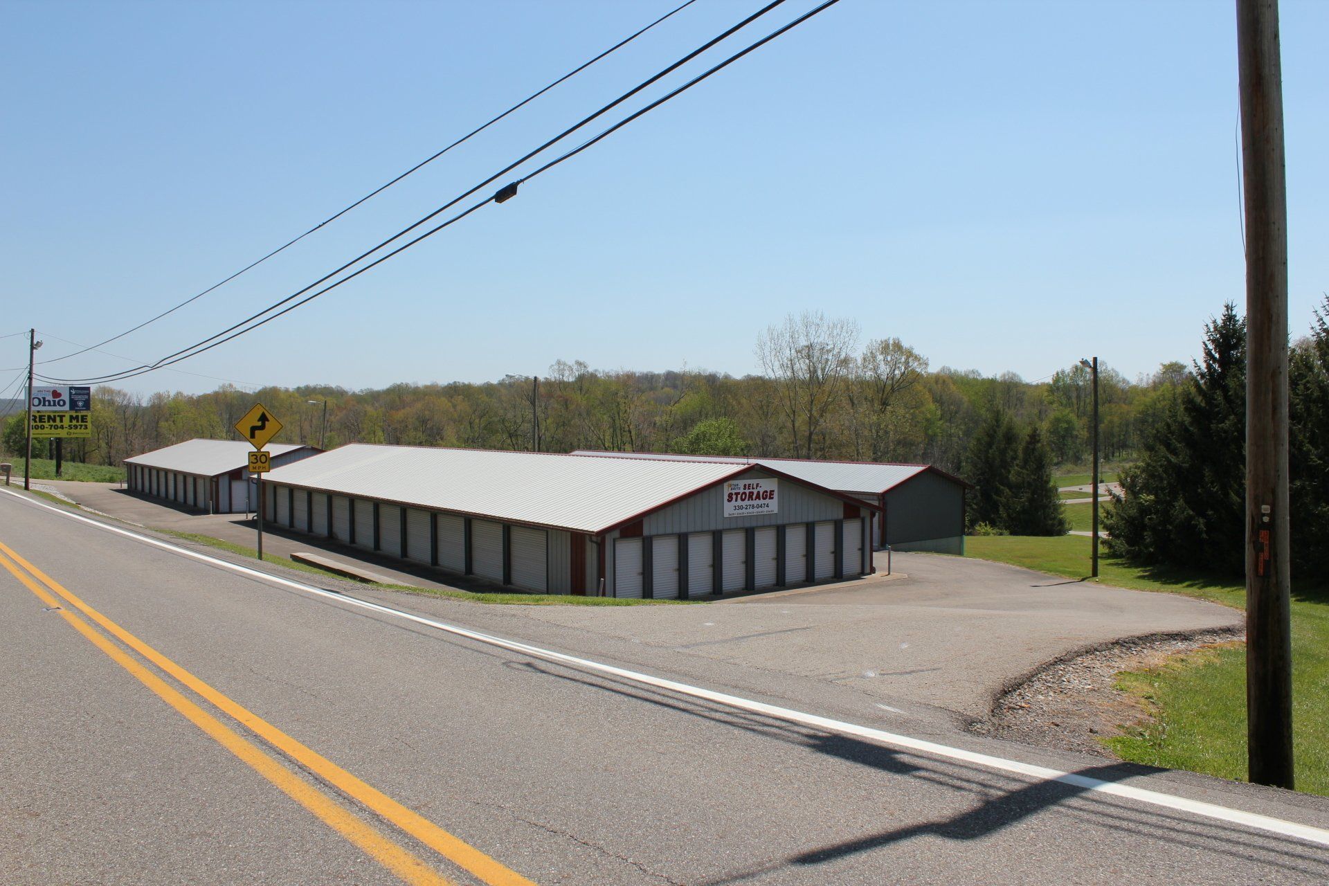 Storage units on the side of a road, silver metal doors, gray roof. Green trees in the background, blue sky.