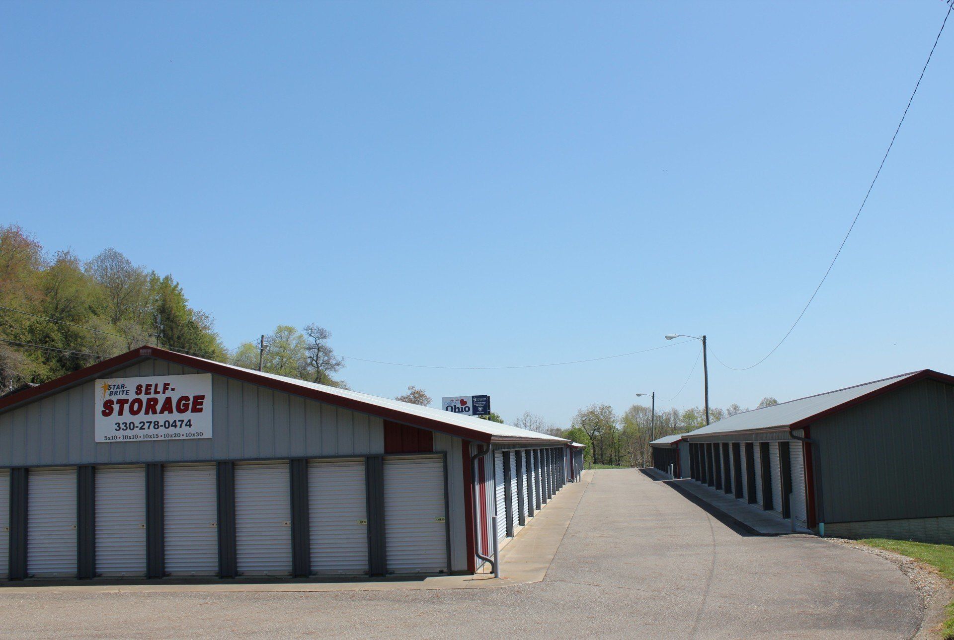 Storage units under a blue sky. Gray buildings with white doors, red roof trim, and asphalt path.