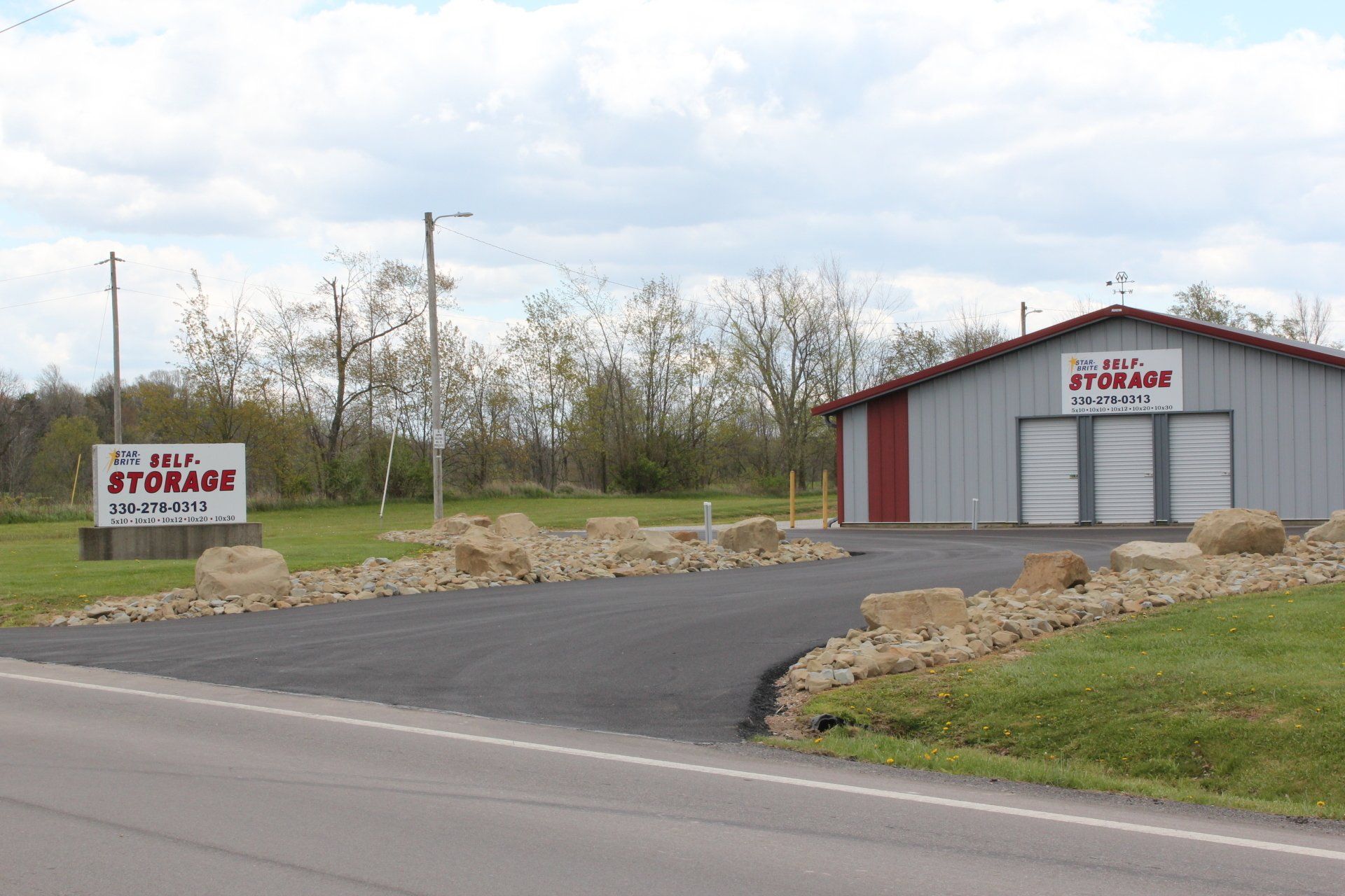 A storage facility with a gray building, red trim, and a sign on a grassy roadside.