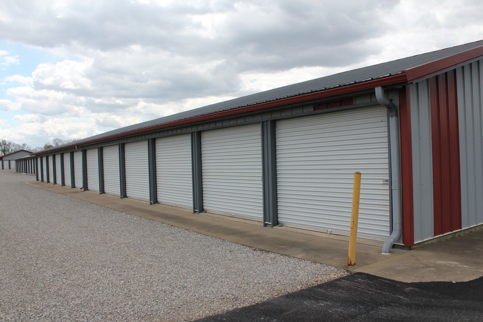 Row of storage units with white doors and gray and red metal siding. Gravel driveway, cloudy sky.