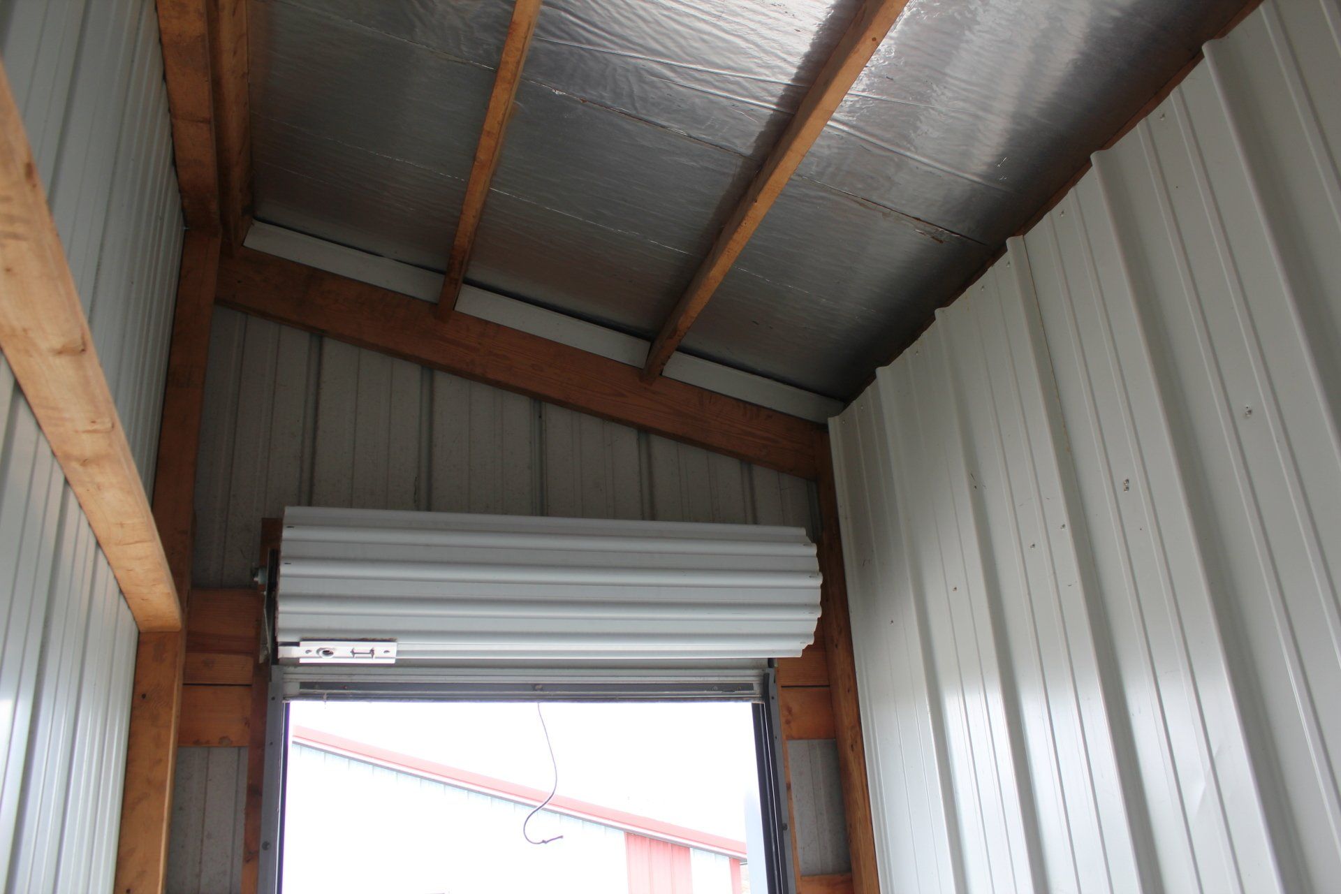 Interior of a metal storage unit with a rolled-up door and corrugated metal walls. Wooden beams support the ceiling.