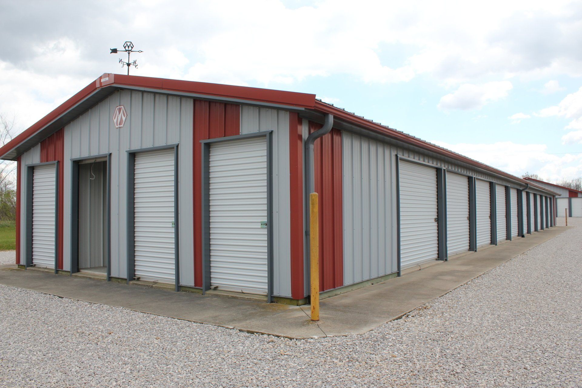 Storage units with red and gray metal siding, roll-up doors, and a gravel base.