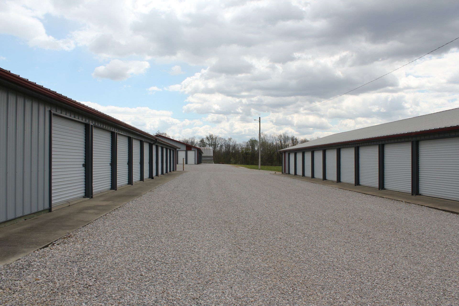 Exterior view of a self-storage facility with rows of gray metal units and a gravel driveway under a cloudy sky.