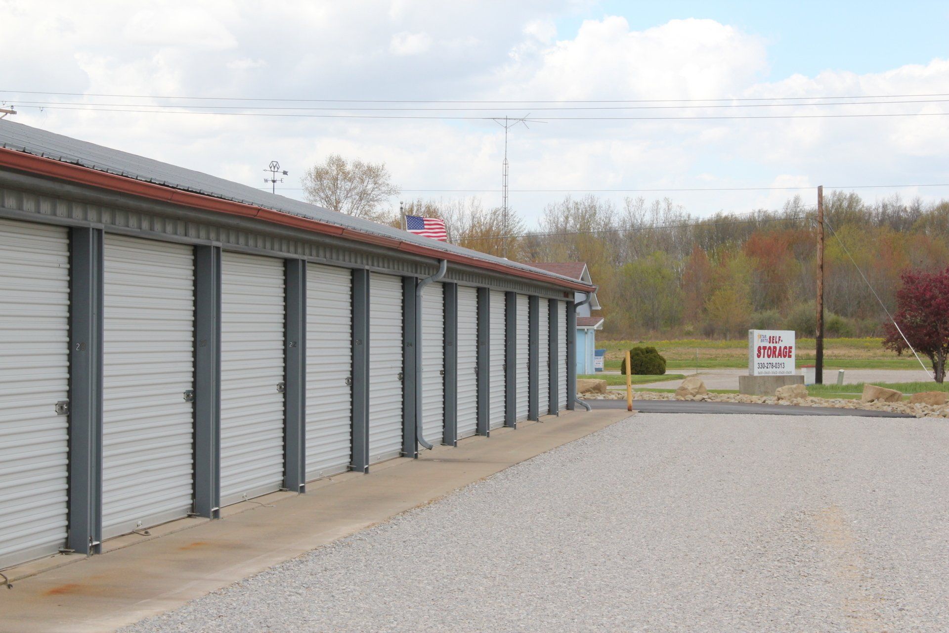 Storage units with white doors, metal frames. Gravel parking lot, overcast sky.